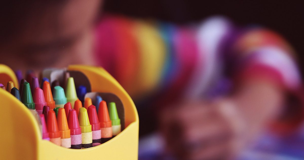 A yellow box of colored crayons in the foreground while a child colors, out of focus, behind it