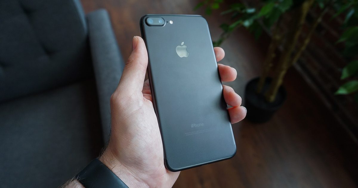 Hand holding a black smartphone face down over a grey sofa, screen hidden from view